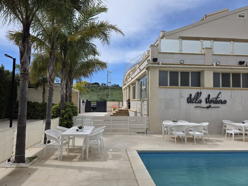 Patio a bordo piscina con tavoli e sedie bianchi accanto a un edificio color crema e palme sotto un cielo azzurro.