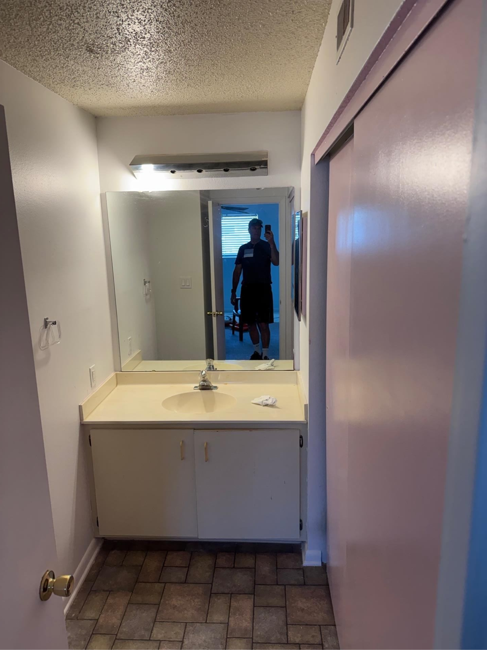 Narrow bathroom with vanity and mirror, seen from the doorway, with a person’s silhouette reflected in the mirror