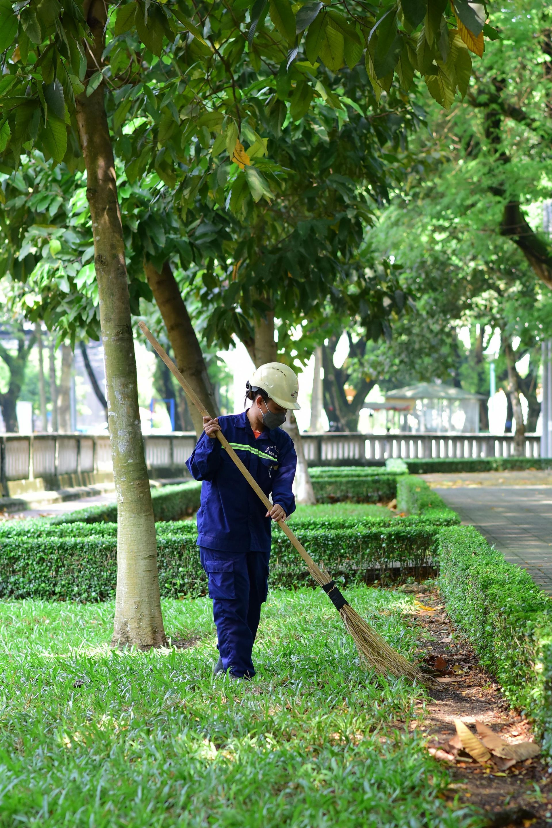 Person in blue uniform sweeping leaves near hedge and trees.