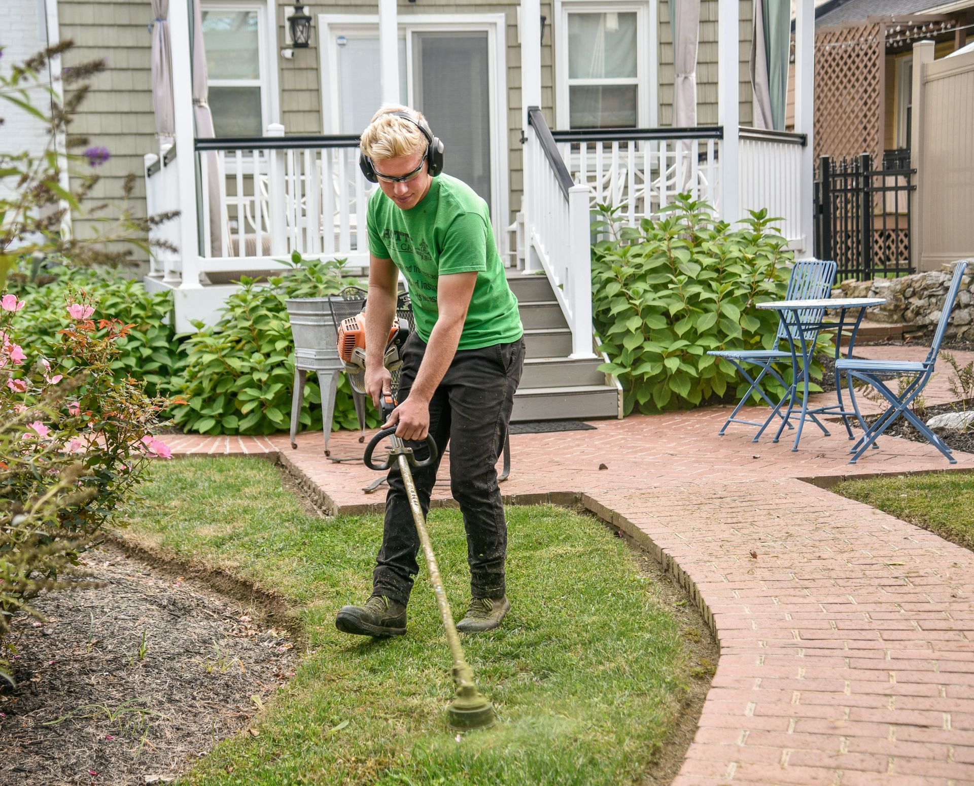 Man using a weed whacker along a brick walkway in a yard, wearing hearing protection.