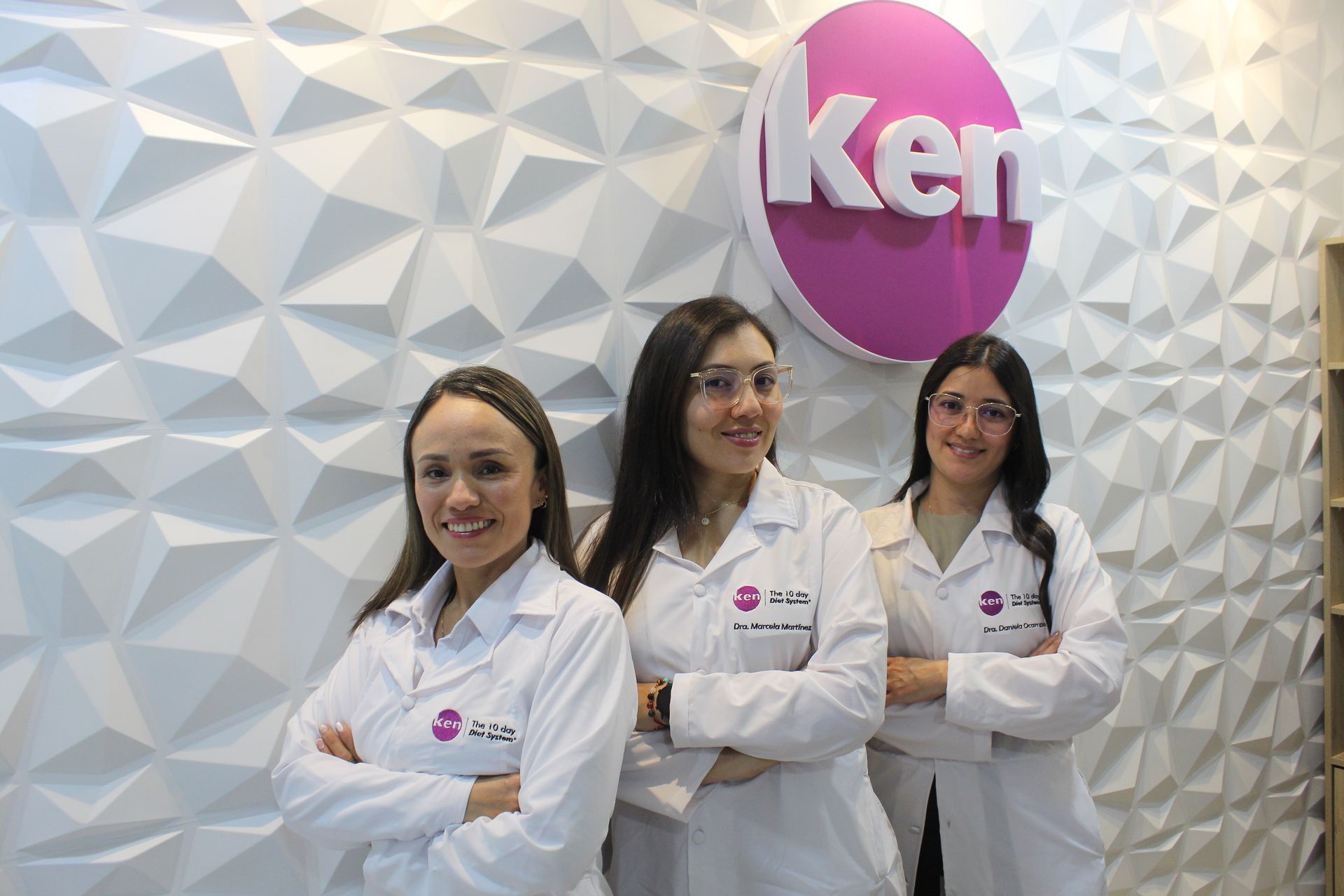 Tres mujeres con batas blancas posan para una foto frente a un cartel de Ken.