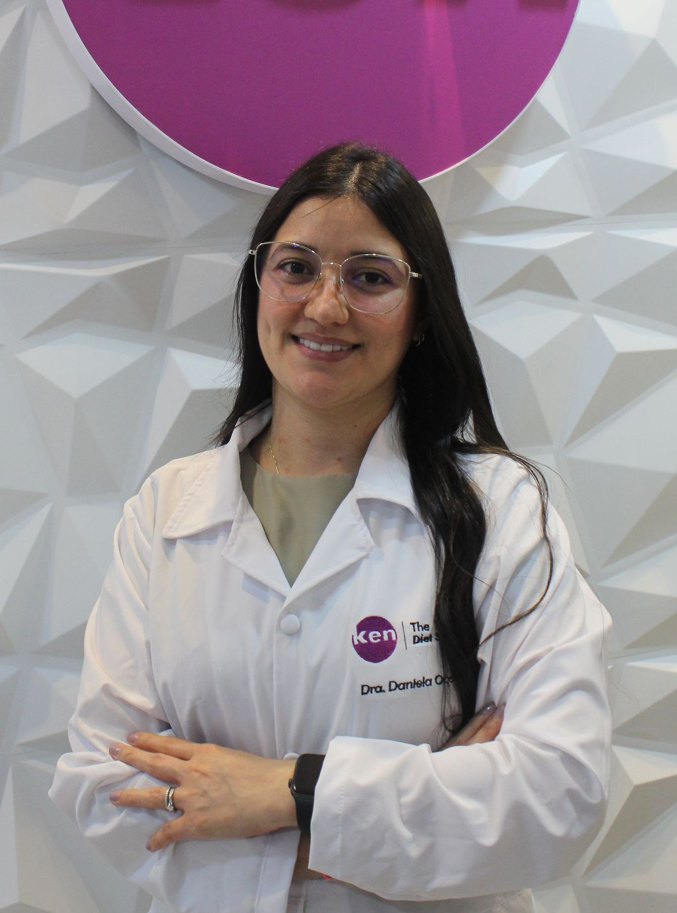 Una mujer con una bata de laboratorio blanca está parada frente a un cartel rosa.