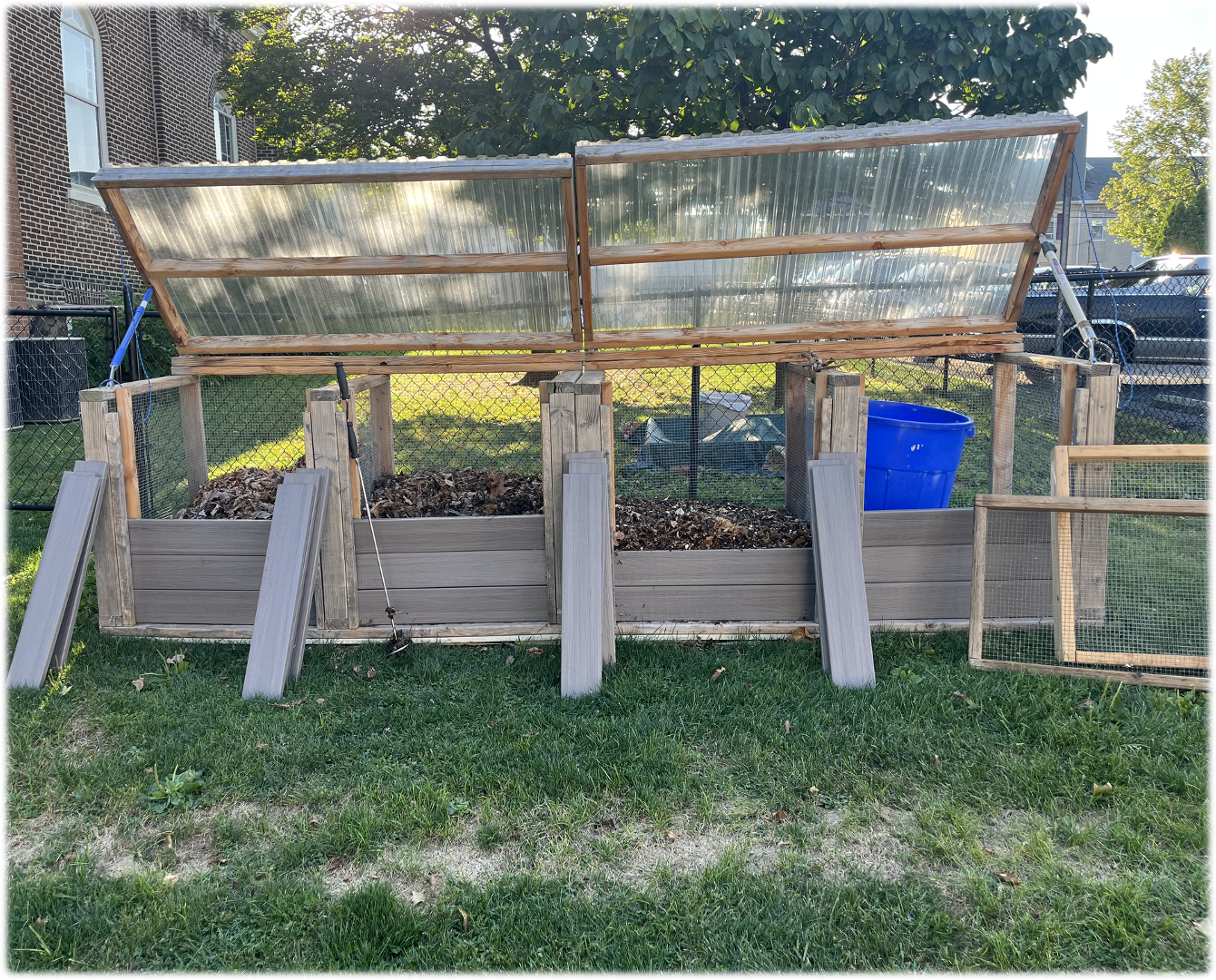 Composting bins with clear, hinged lids made from wood and corrugated plastic, on green grass.