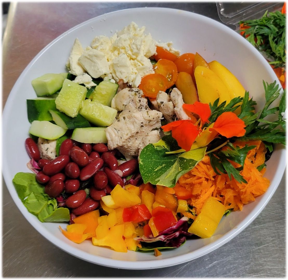Salad bowl with various ingredients: beans, cheese, chicken, tomatoes, cucumber, carrots, mango, edible flowers.