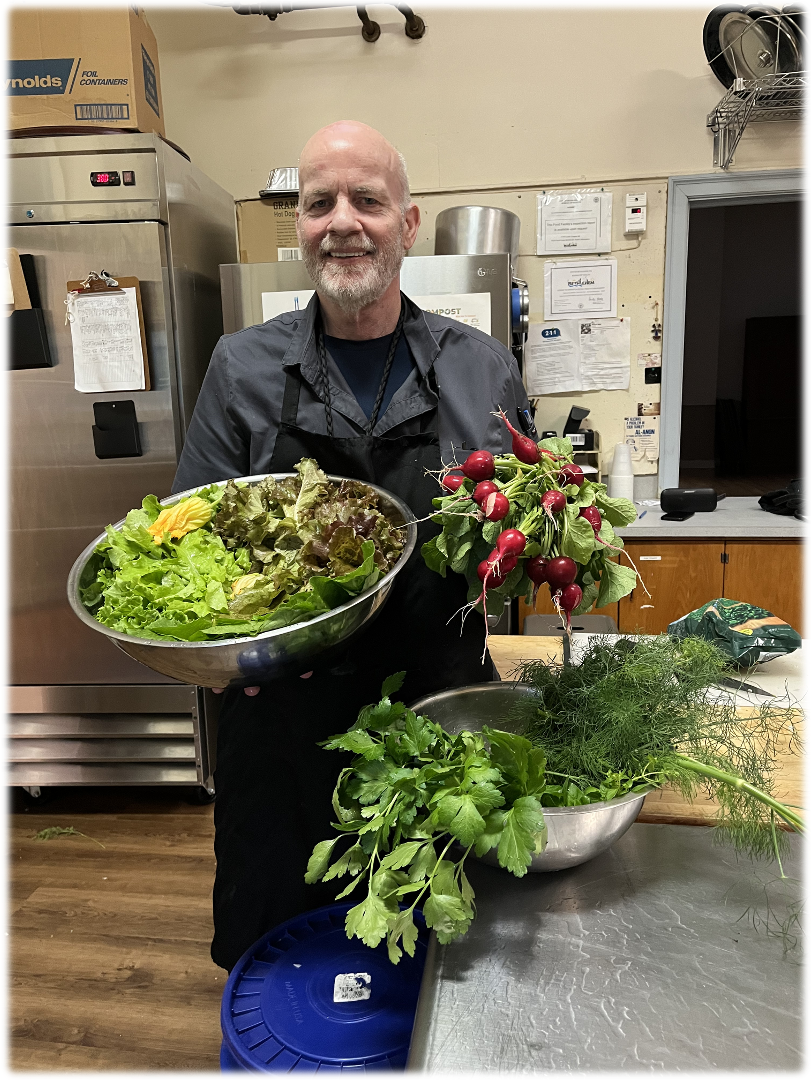 Man in a kitchen holding bowls of fresh produce: lettuce, radishes, and cilantro.