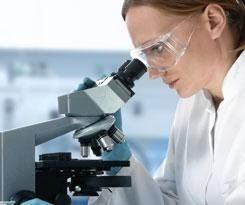 Donna in camice e occhiali protettivi che osserva un microscopio in un laboratorio.