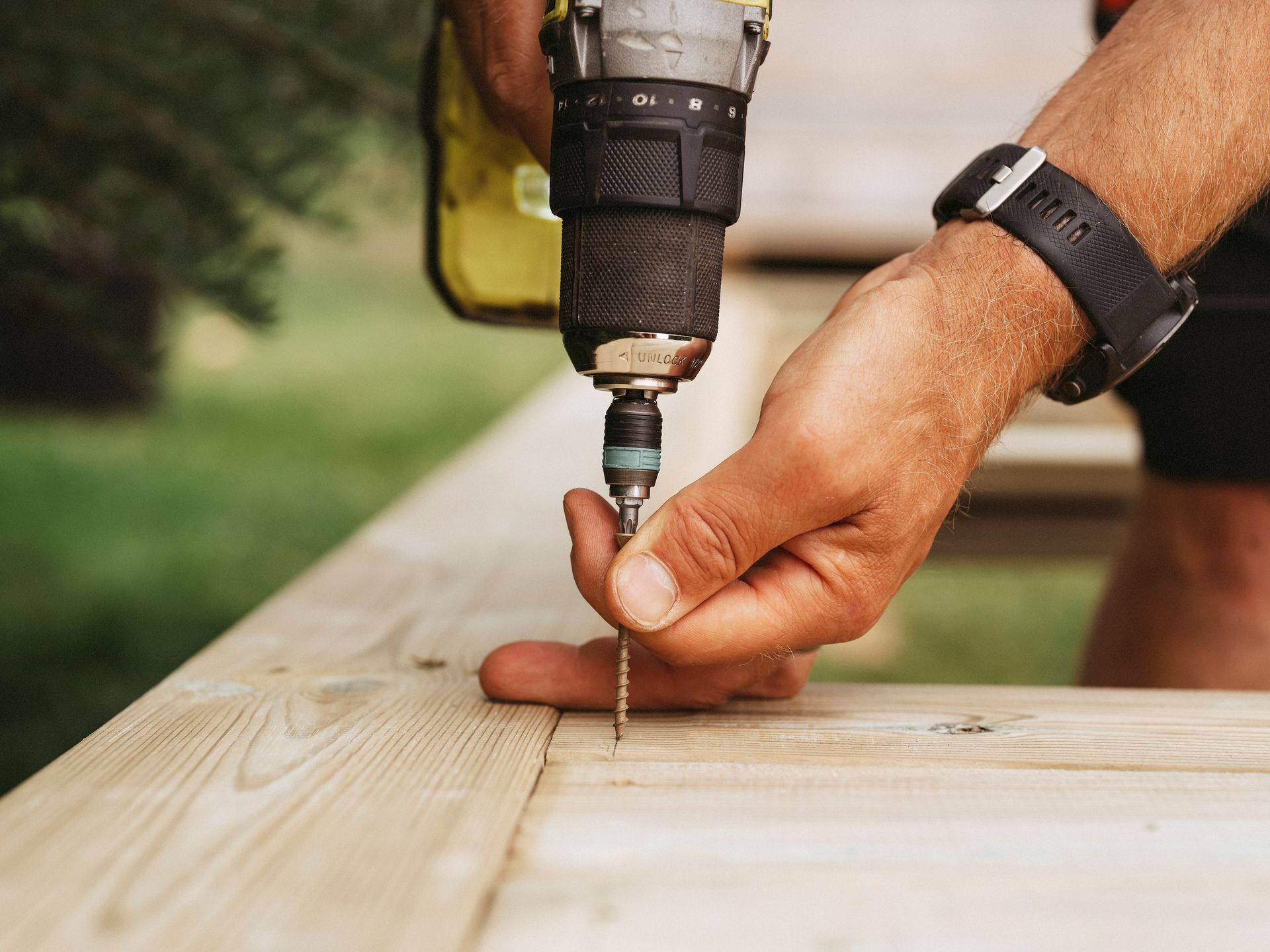 Person using a drill to screw into a wooden surface outdoors.