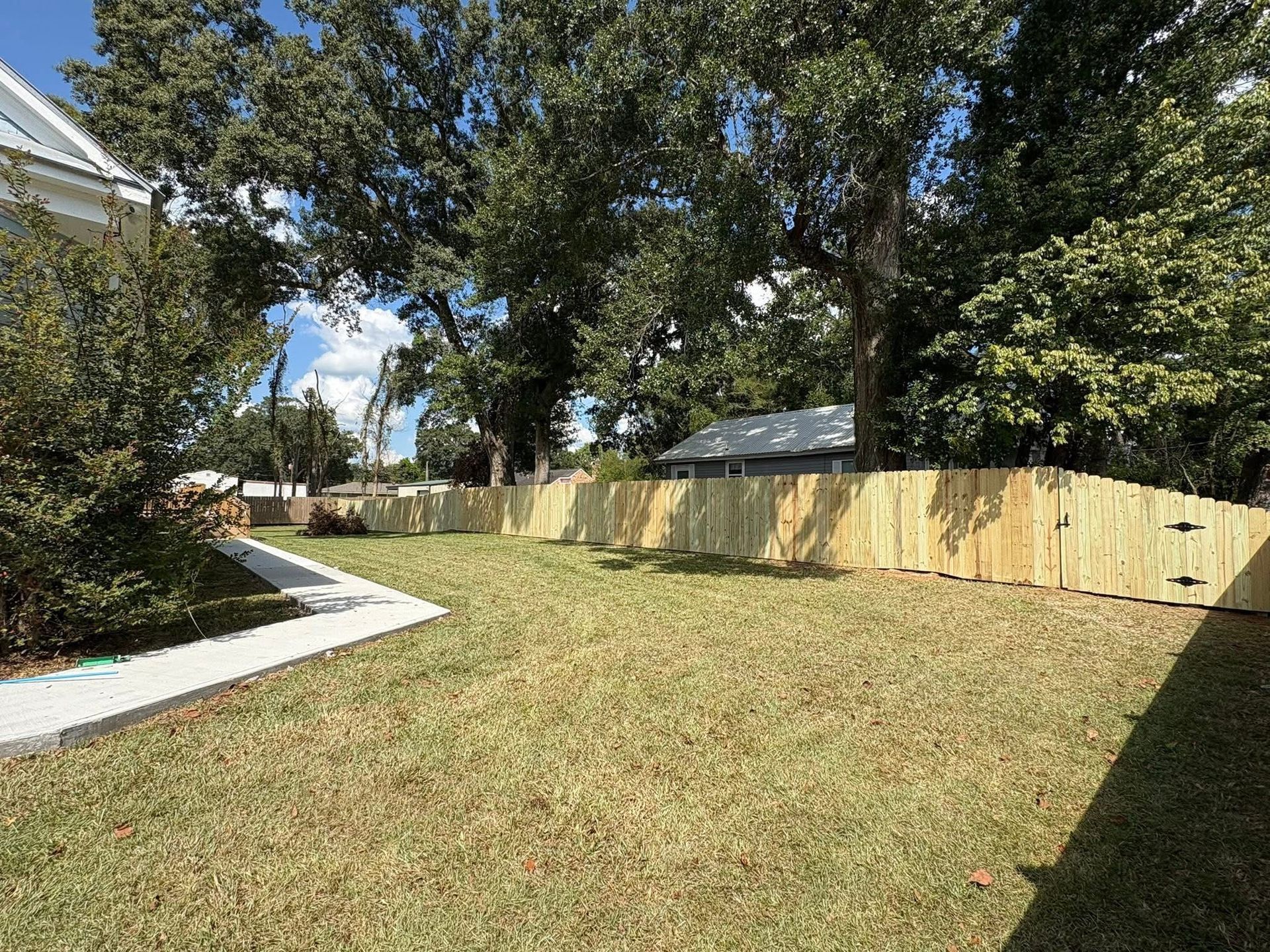 Wooden fence enclosing a grassy yard under trees on a sunny day. A building is visible behind the fence.
