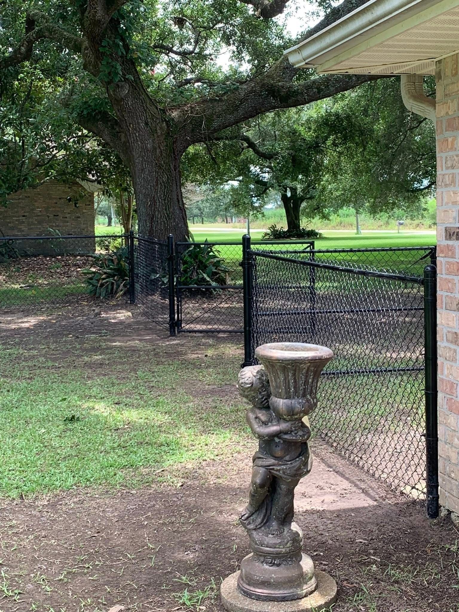 Statue of a cherub holding a flower pot in front of a wrought iron fence, a tree, and a grassy area.