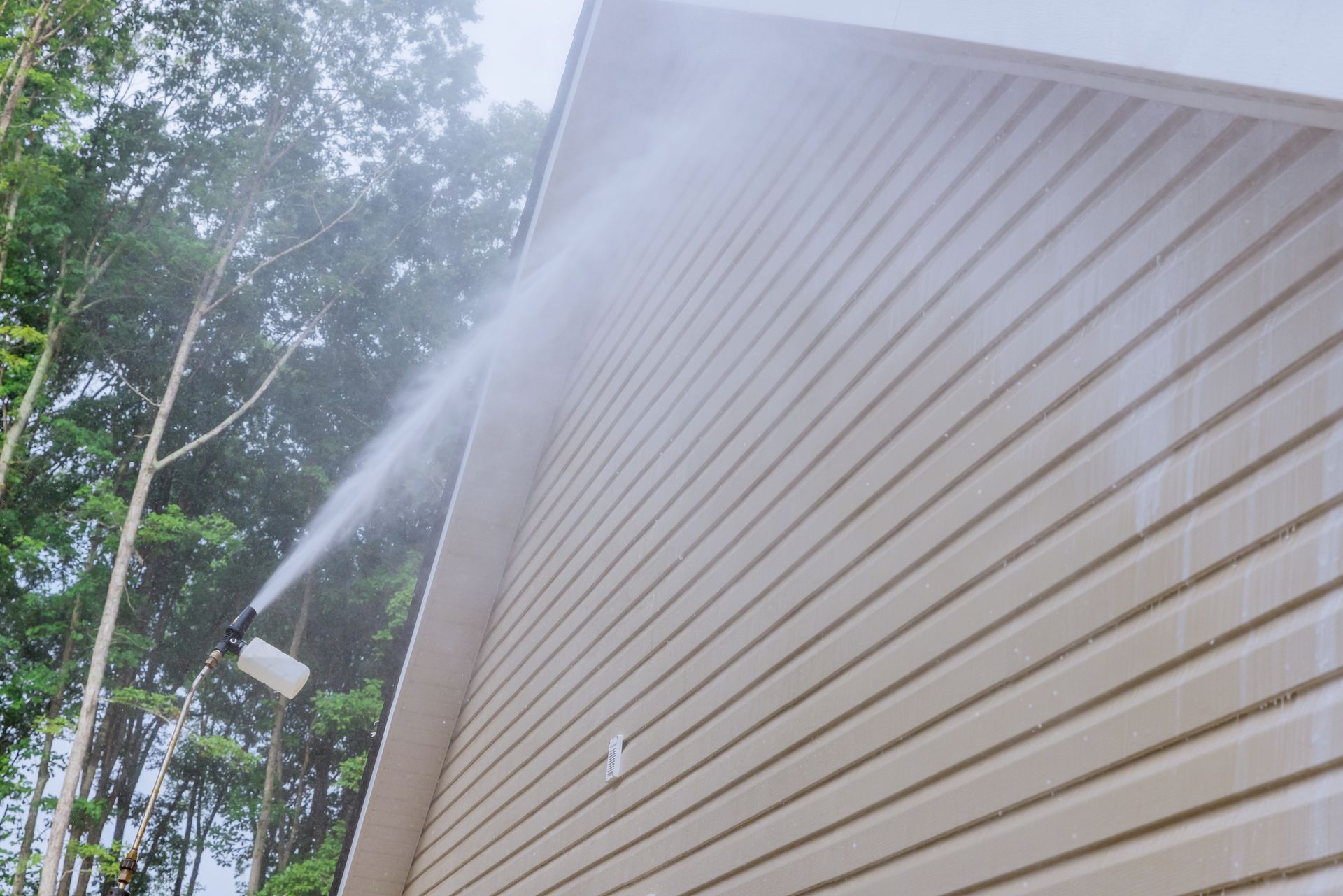 A high-pressure nozzle sprays a stream of cleaning solution onto the light-colored vinyl siding of a house.