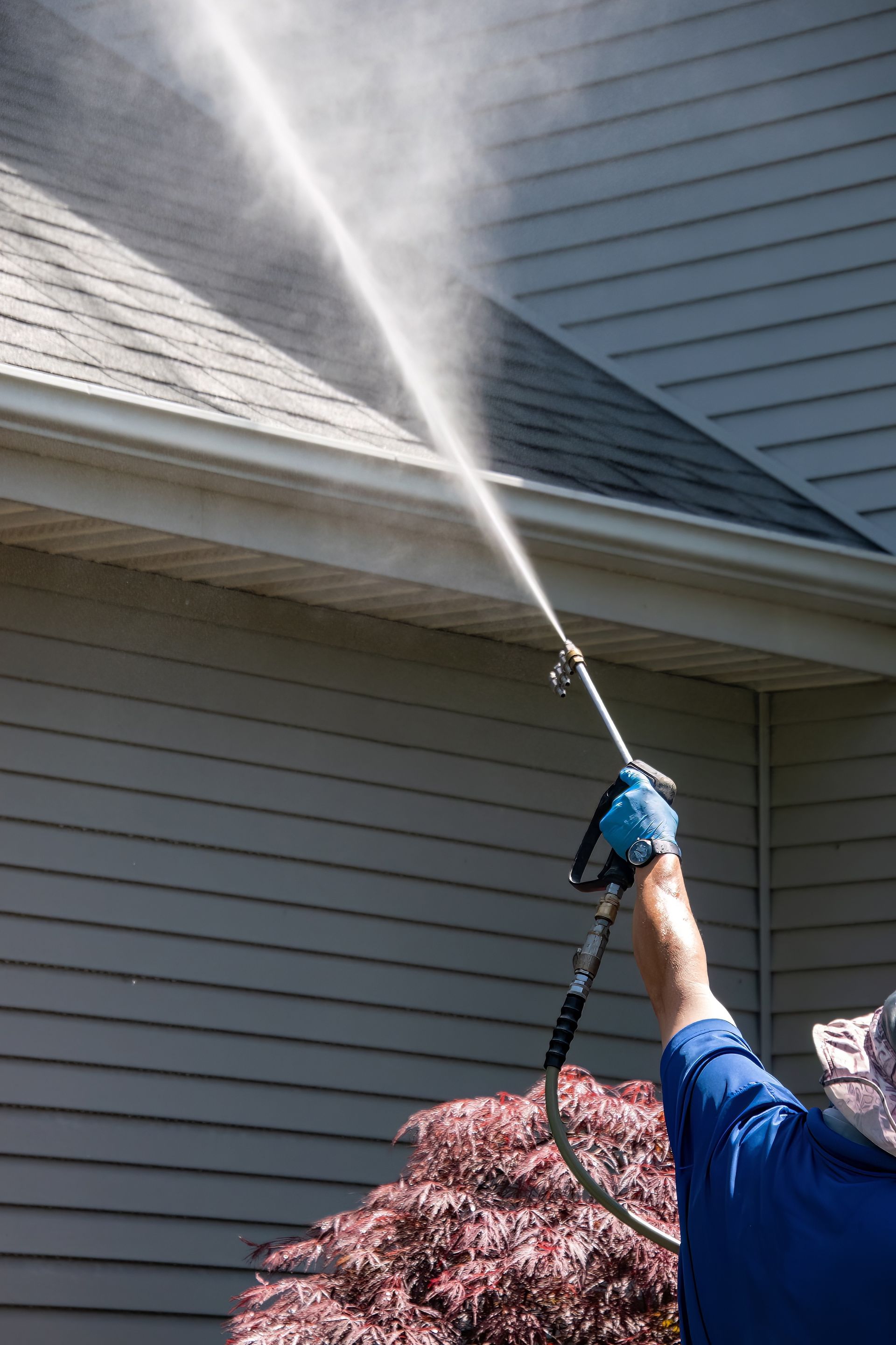 A person uses a pressure washer to clean a shingled roof on a house with grey siding.