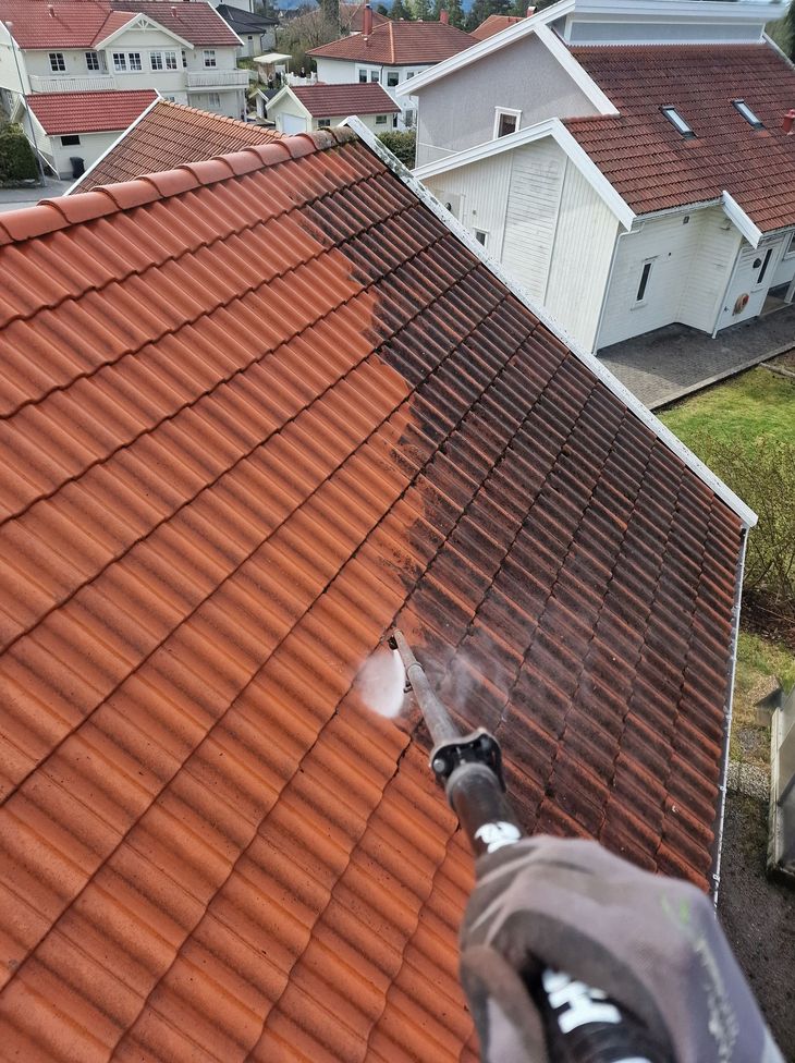 A first-person view of a gloved hand pressure washing a dark, dirty roof tile section, revealing clean, red tiles underneath.