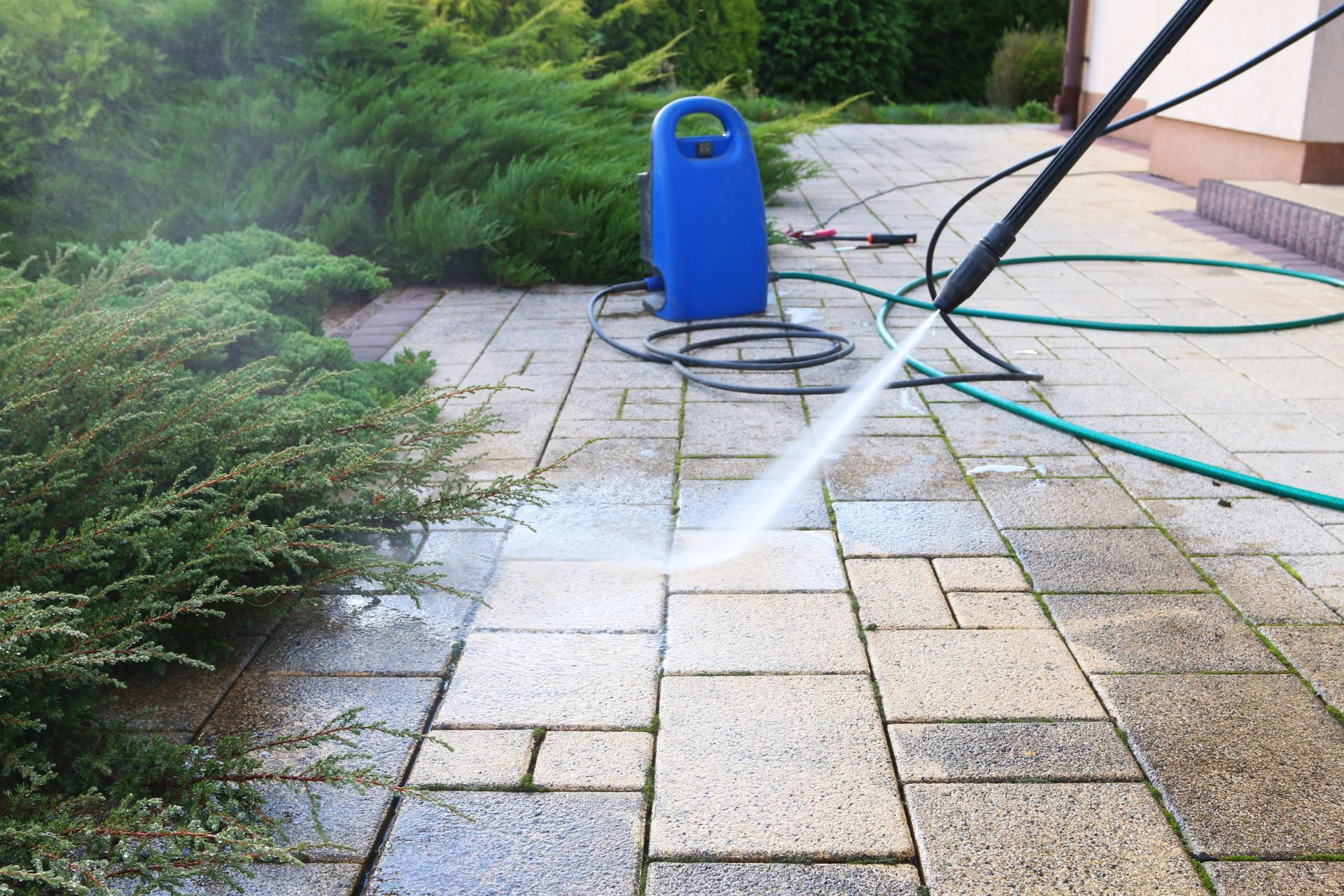 A high-pressure washer sprays water onto light-colored paving stones near a blue machine and green garden shrubs.