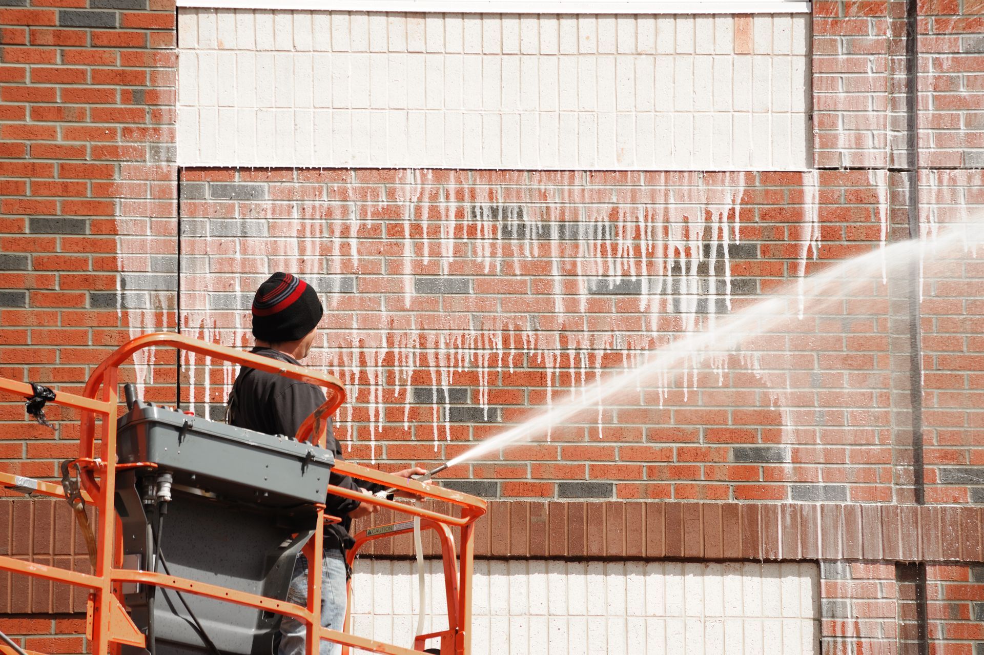 A person in a lift power washes a red brick building, spraying white liquid across the wall.