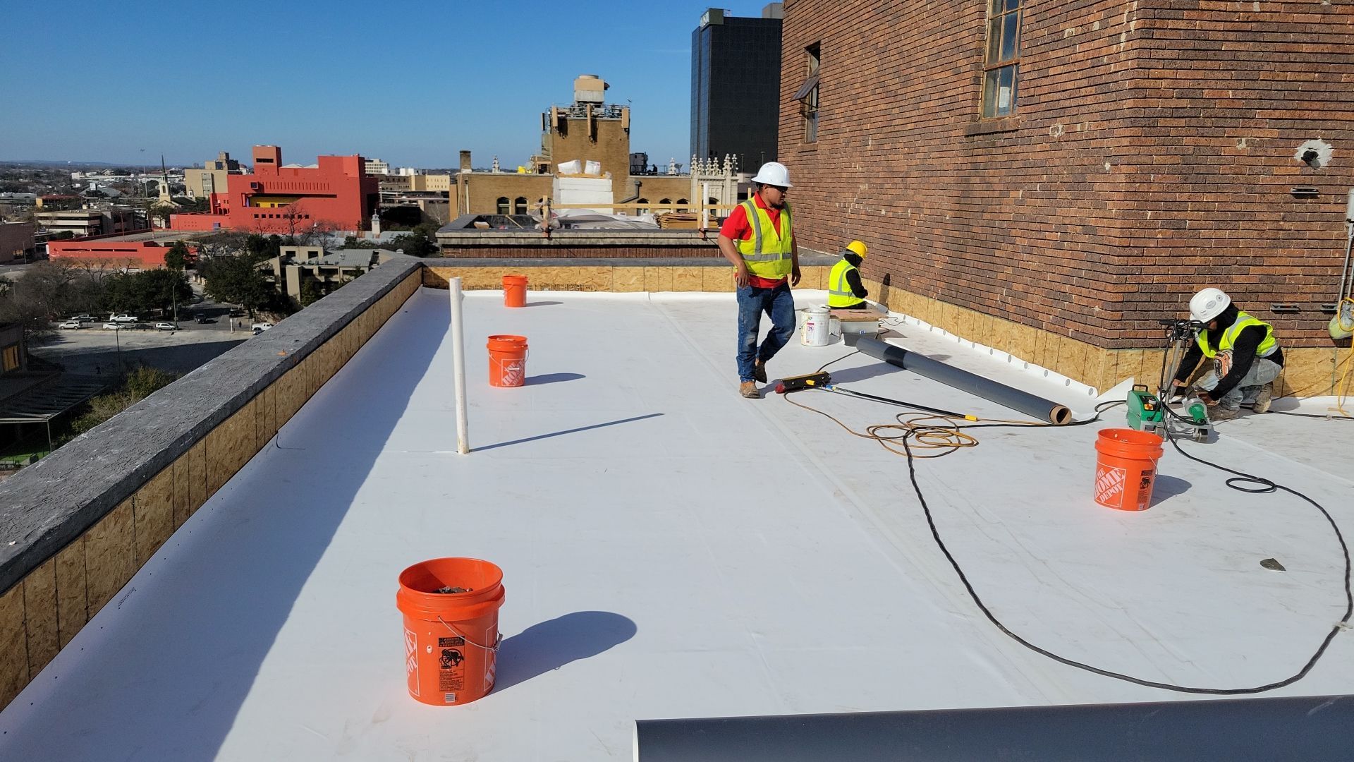 Construction workers on a flat roof, installing white roofing material. Orange buckets, brick building, city background.
