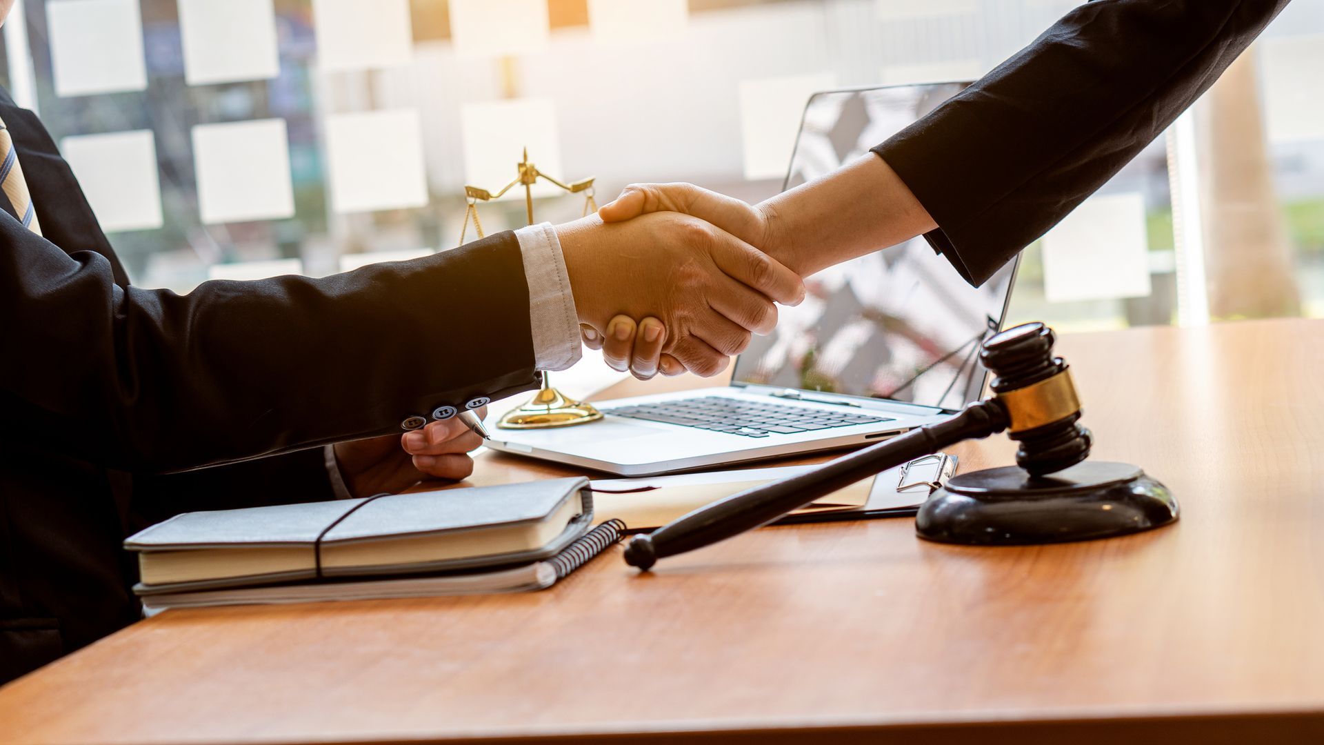 Two people shaking hands over a table with a gavel, legal books, and a laptop.