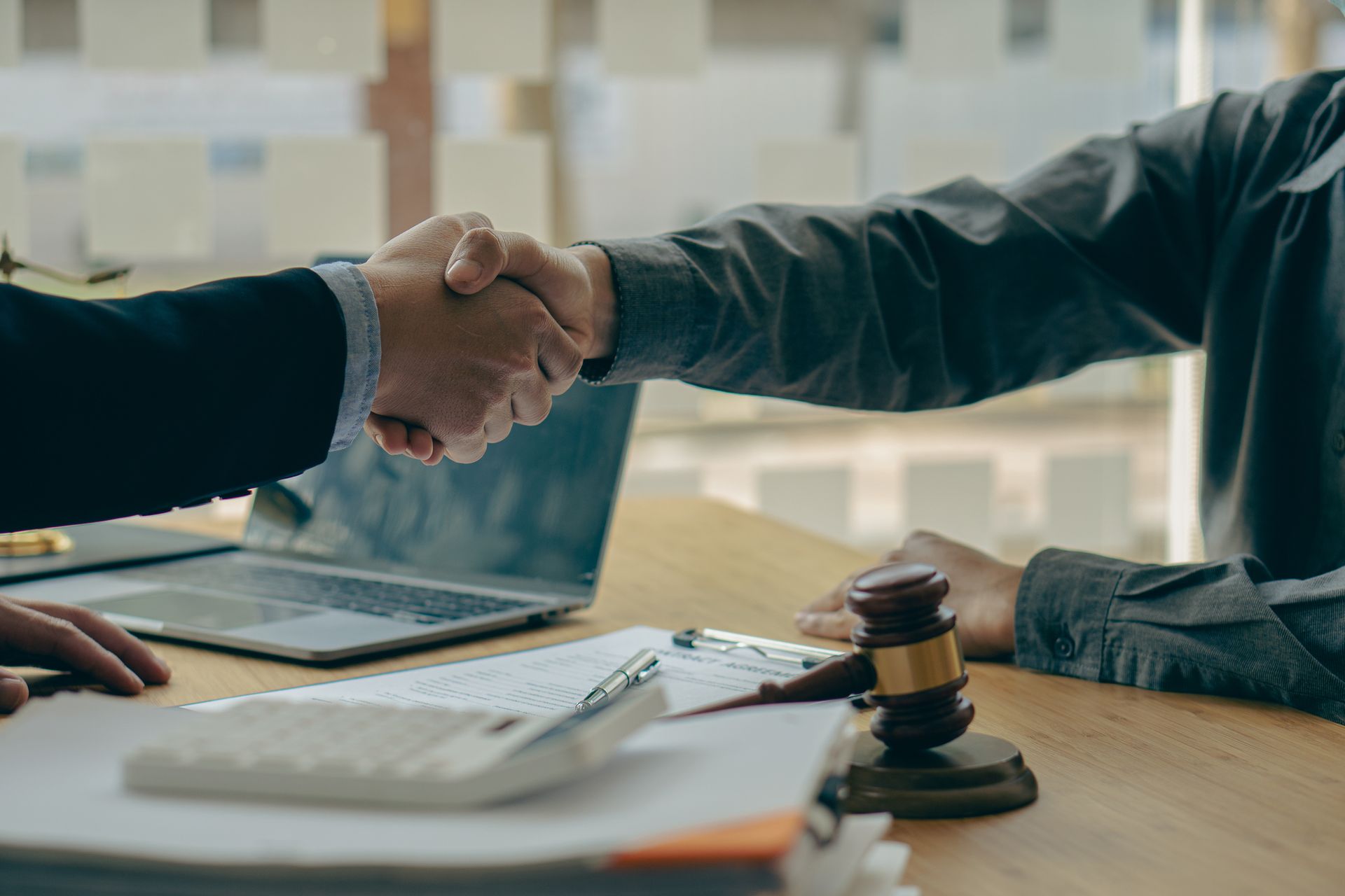 Two people shaking hands over a desk with documents, a gavel, and a laptop.