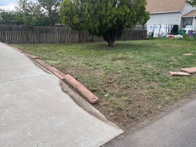 A concrete driveway leading to a yard with a tree and a fence.
