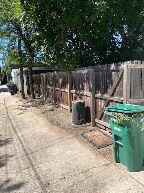 A green trash can is sitting on the sidewalk next to a wooden fence.