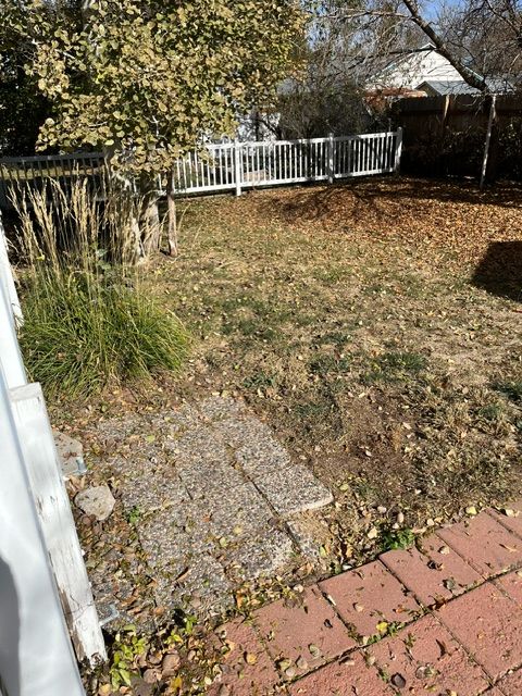 A backyard with a brick walkway and a white fence.