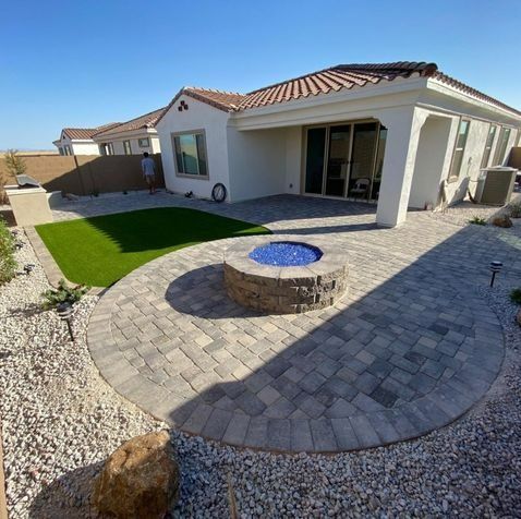 Backyard patio with fire pit, artificial turf, and a house under a blue sky.