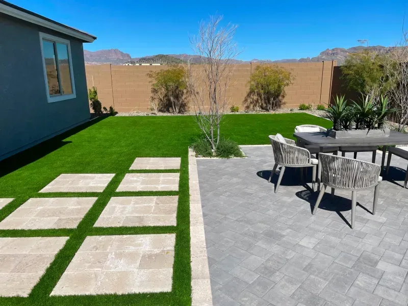 Backyard with patio furniture, green lawn, square pavers, and beige wall under a blue sky.