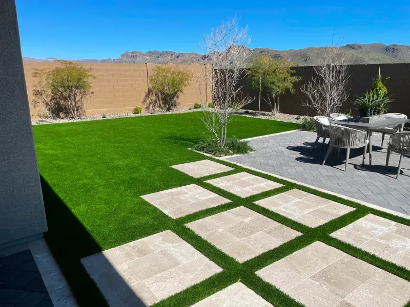 Backyard with green turf, stone patio, and desert landscape under a blue sky.