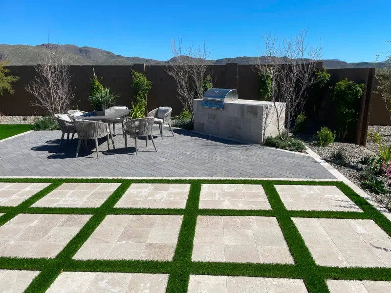 Patio with outdoor kitchen, dining set, and grass squares in a desert landscape.