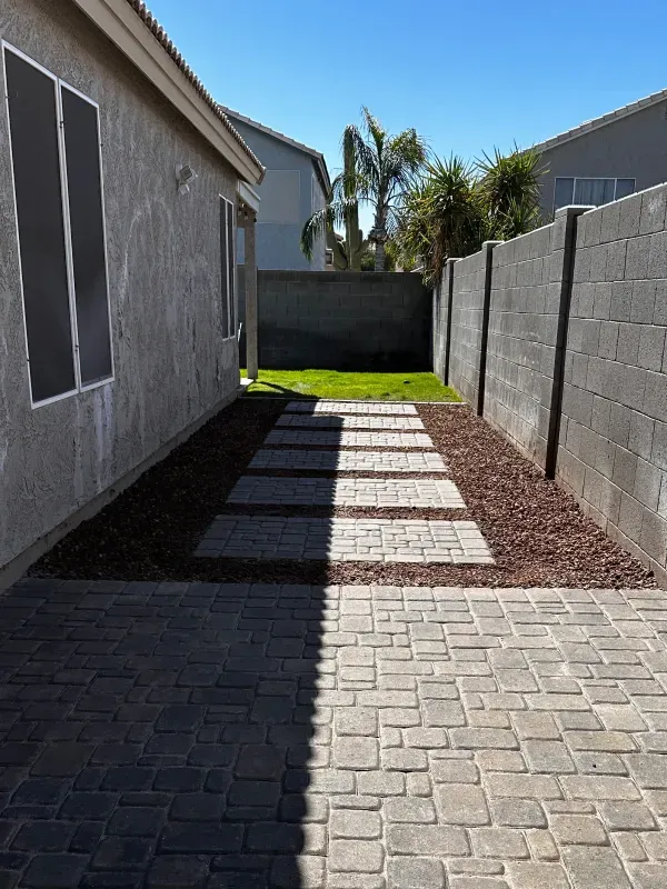 A narrow backyard with a stone walkway, gravel, and a brick wall. Bright sunlight.