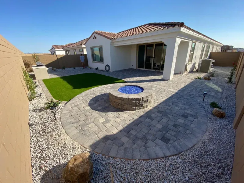 Backyard patio with fire pit, paved area, artificial turf, white stucco house, and gravel landscaping.