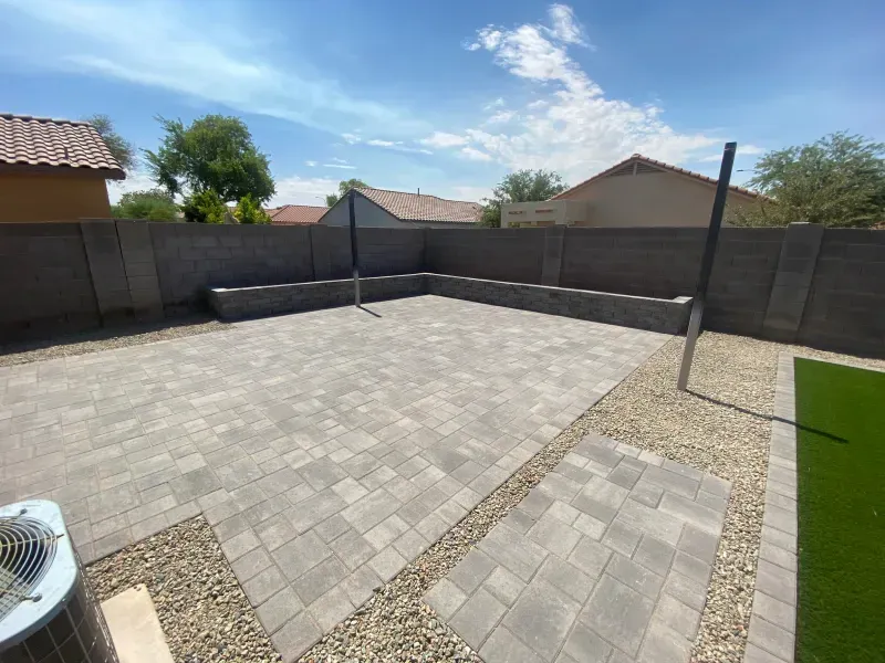 Backyard patio with pavers, rock border, and artificial turf, enclosed by a wall on a sunny day.