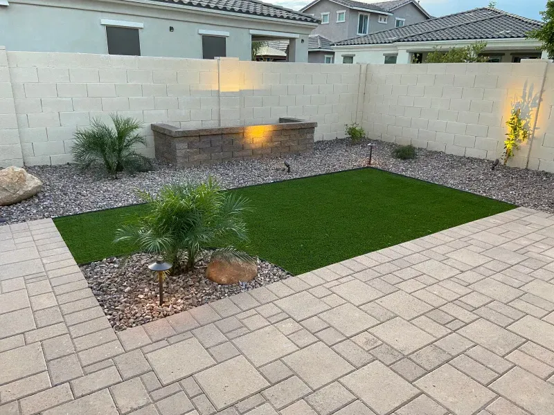 Backyard with artificial turf, gravel, stone pavers, and decorative walls, plants and lighting.