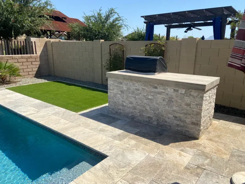Poolside outdoor kitchen with stone counter and grill. Turf, tan walls, and blue water.