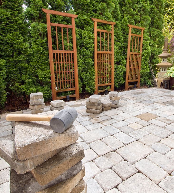 Patio with pavers under construction, mallet, stacked blocks, wooden arbors, and lush green hedge.