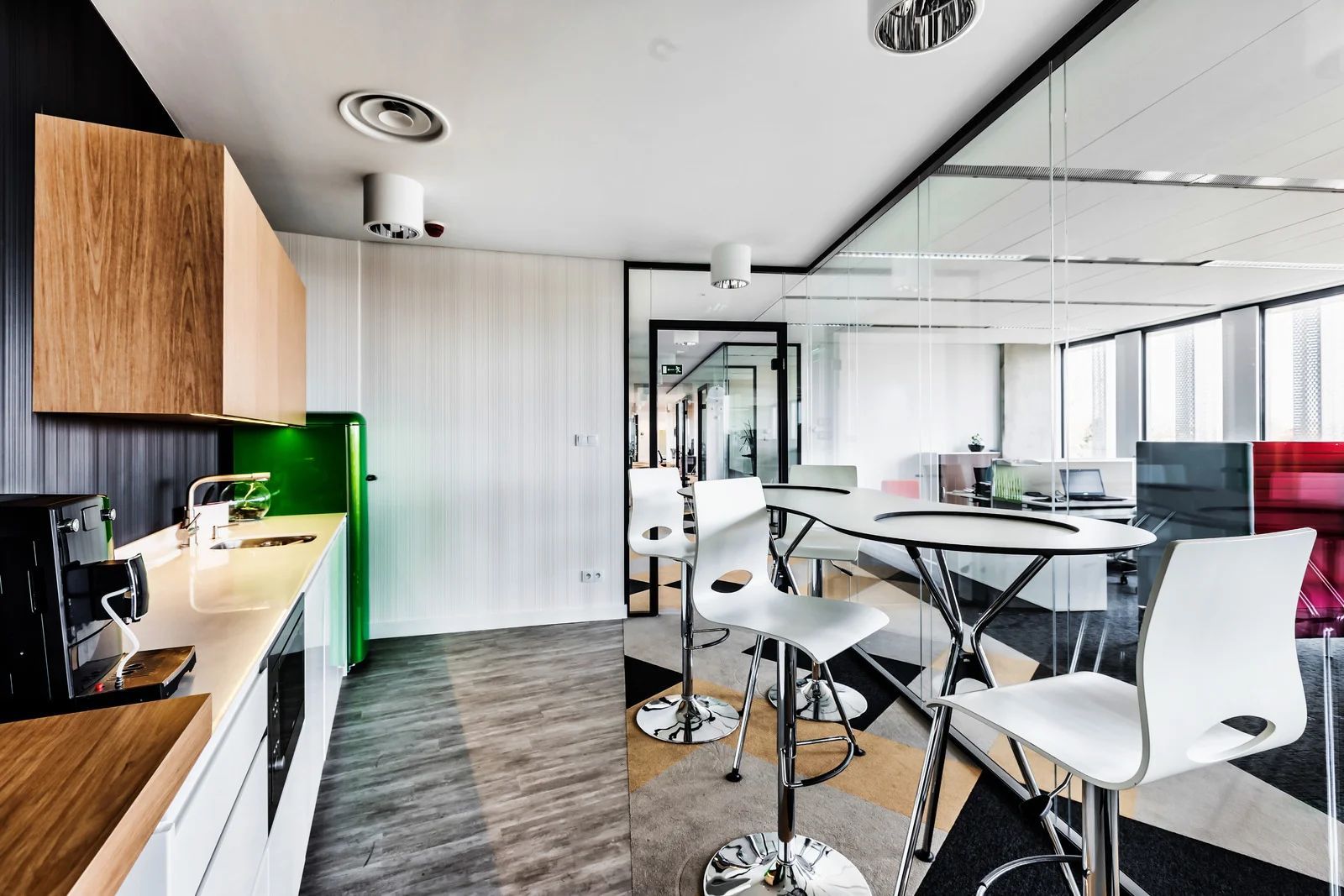 Modern office kitchen with a white countertop, wooden cabinets, and green refrigerator. White chairs surround a table.