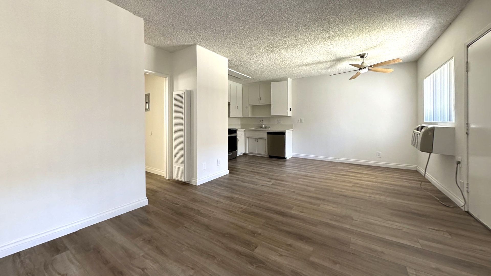 Empty, bright room with wood-look floor, white walls, and a partial view of a kitchen.