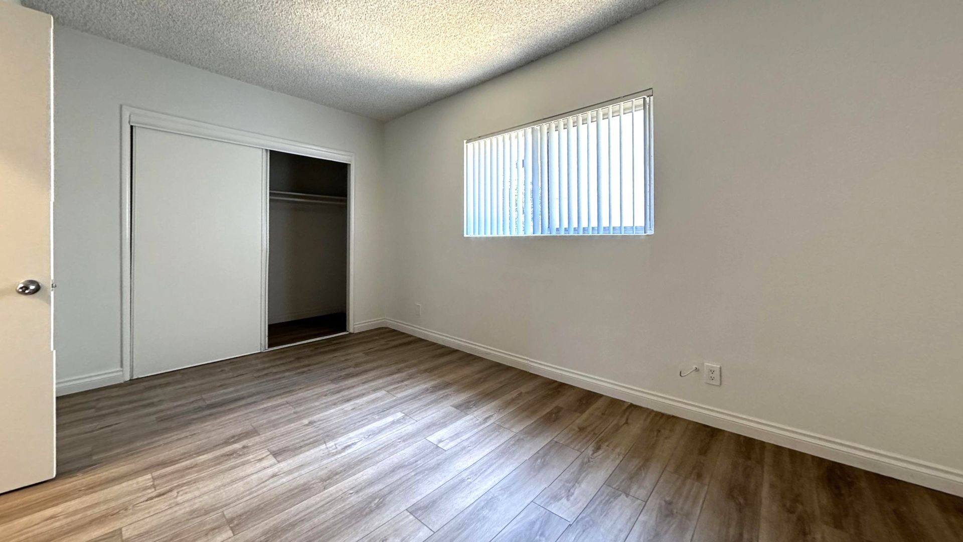 Empty bedroom with closet, window, and wood-look flooring. White walls and ceiling.