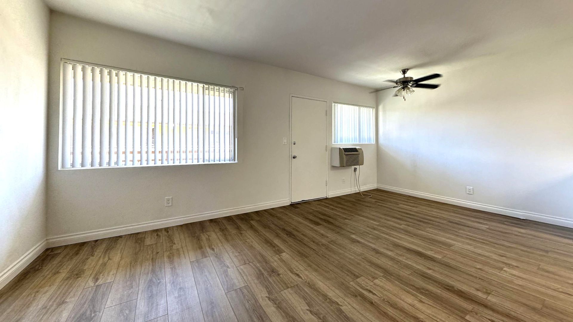 Empty room with wood-look flooring, white walls, and a window with blinds. A door and an air conditioner are visible.