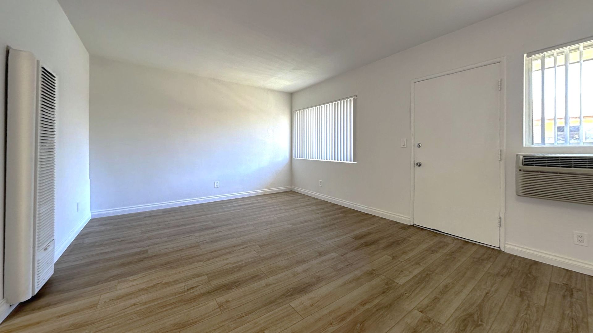 Empty room with wood-look flooring, white walls, two windows, and a door. An air conditioner is in one window.