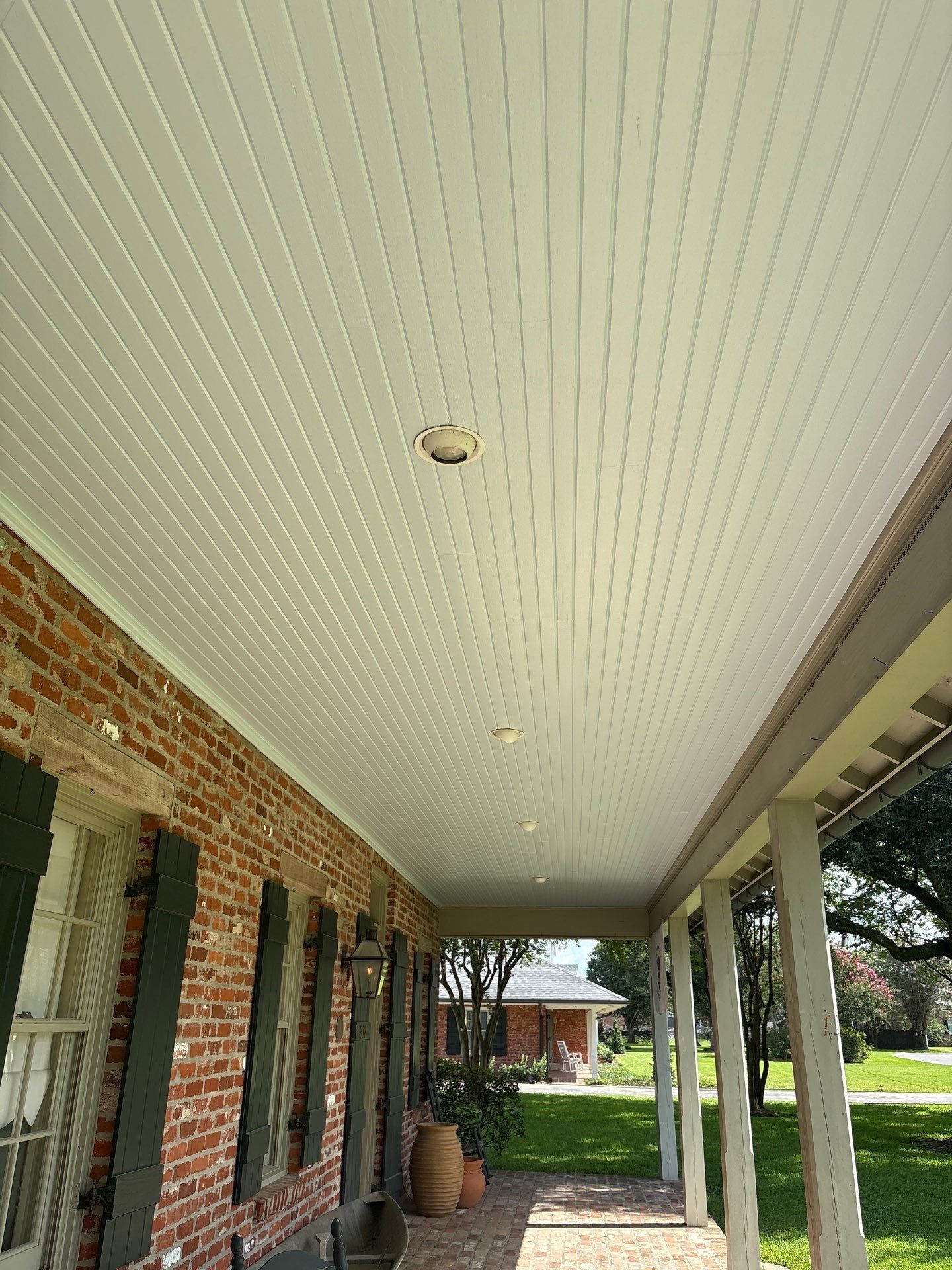 A long porch with a white-paneled ceiling. Brick wall on the left, pillars on the right, and trees in the background.