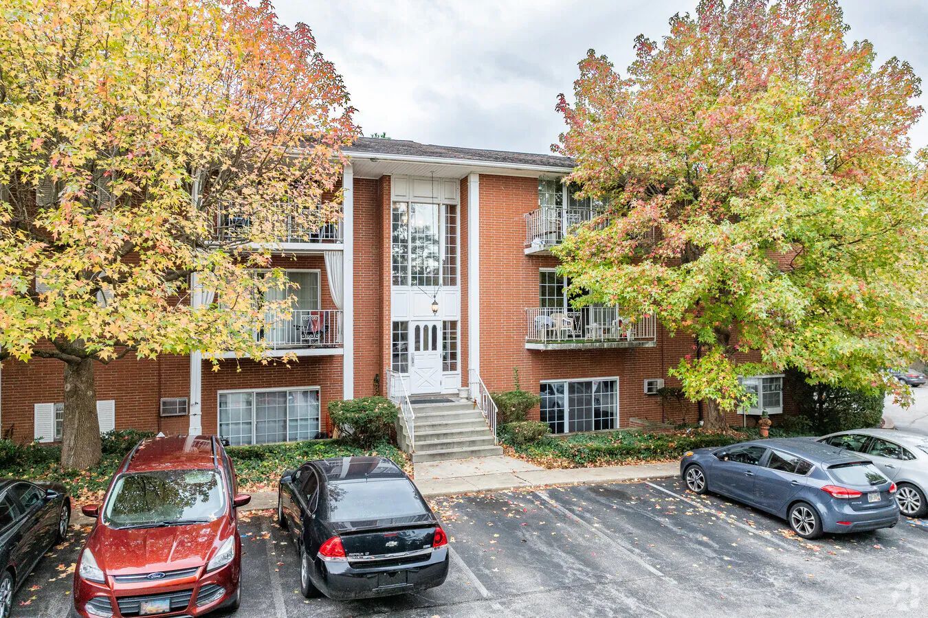 Apartment building with brick exterior, three stories, small balconies, autumn trees, and parked cars.