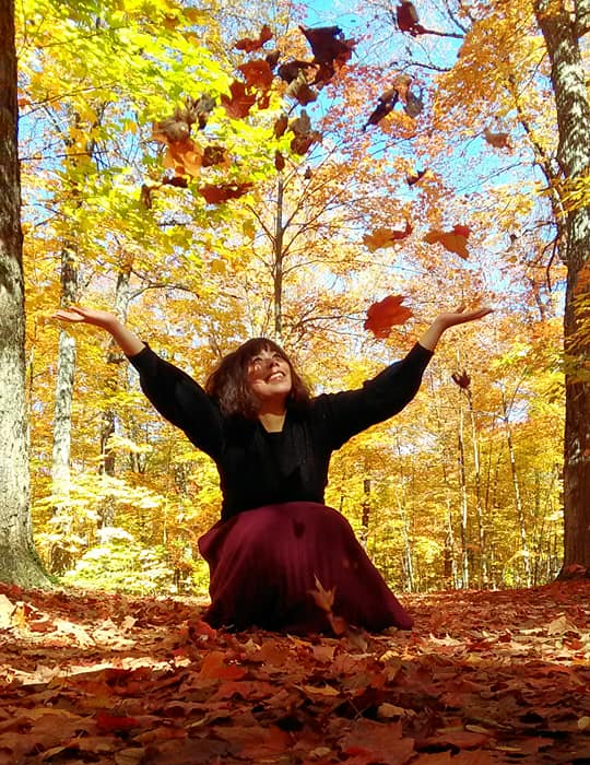 A woman kneeling down with arms open and looking up at the fall leaves she's thrown overhead.