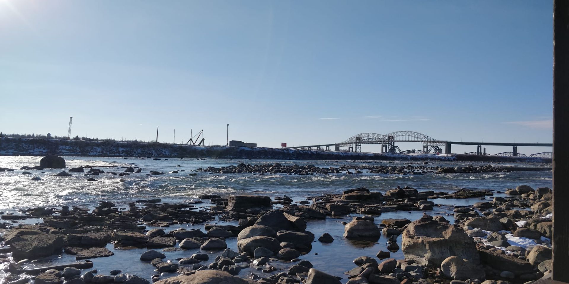 An image of the Sault Bridge that connects Canada to Michigan on a cold winter day. The sky is blue and the shores are rocky.