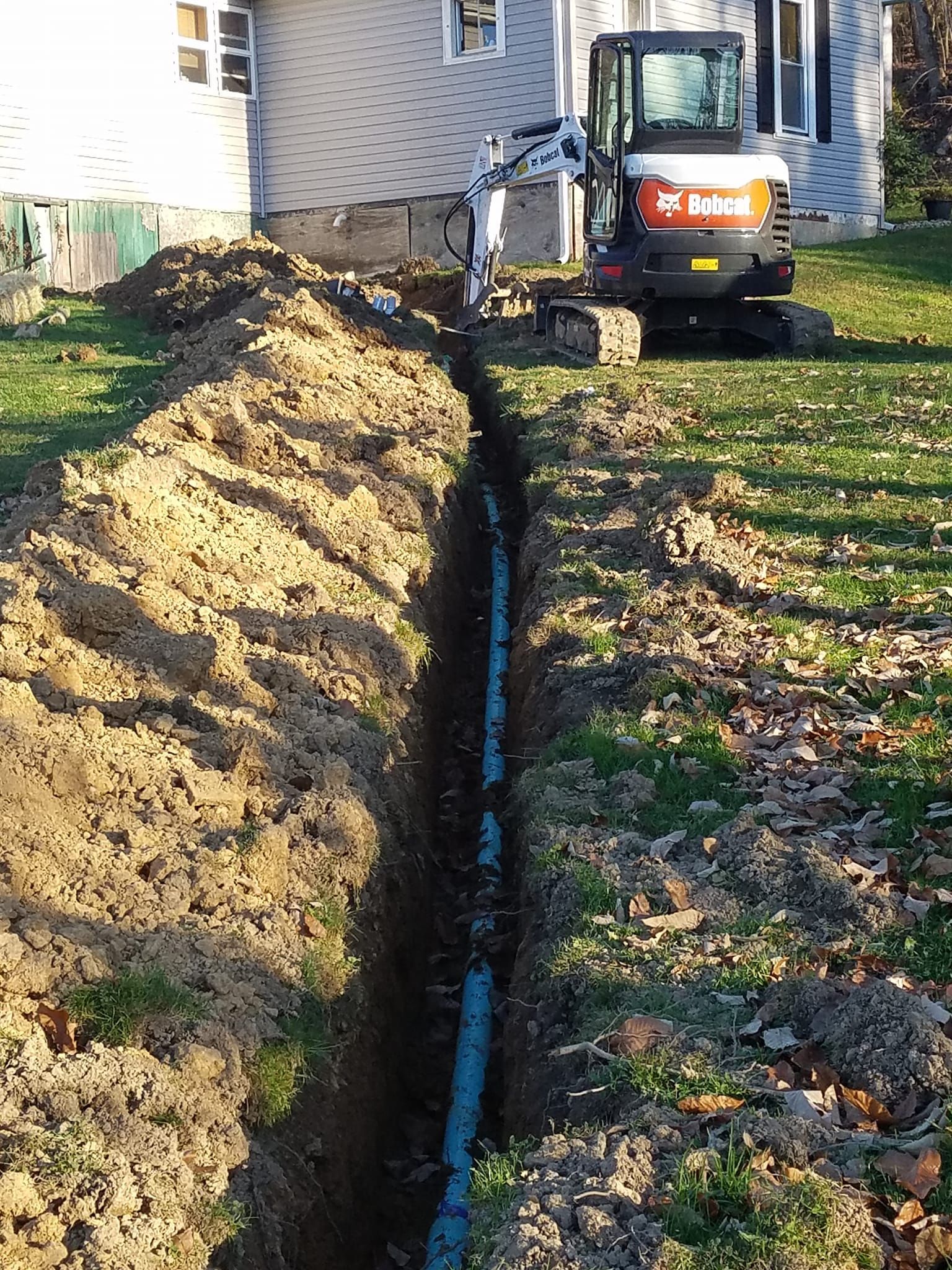 Bobcat excavator digging a trench, with buried pipe visible, beside a house on a lawn.