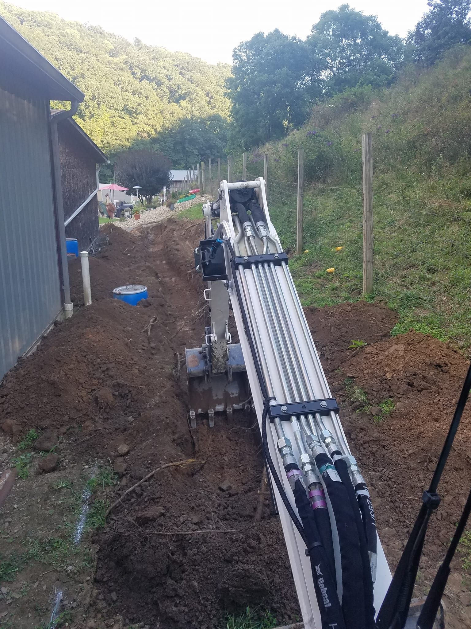 Excavator digging a trench next to a building and hillside in an outdoor setting.