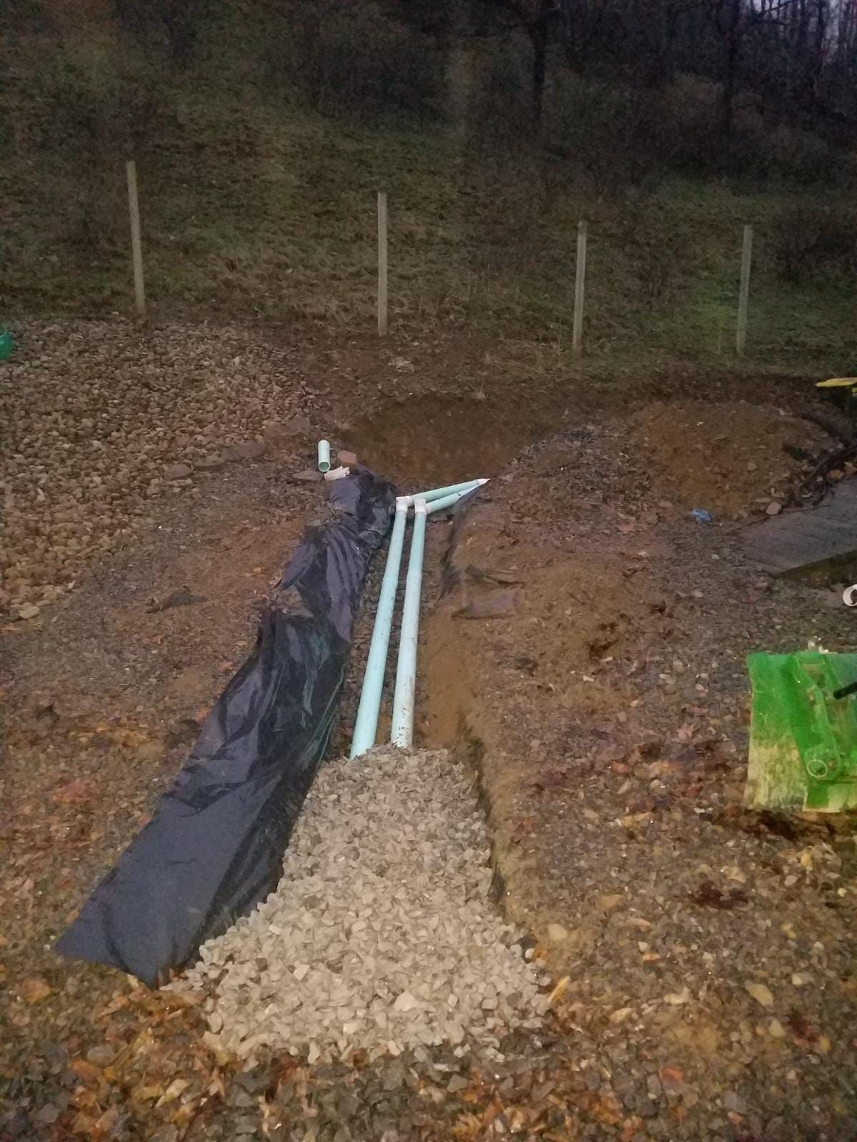 Trench with buried blue pipes, gravel, and black fabric in a hillside for drainage.