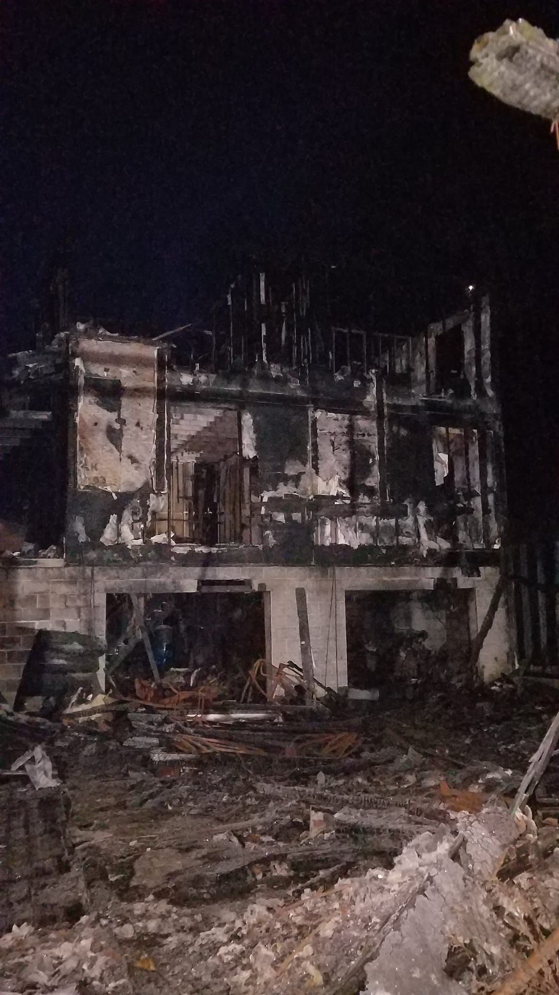 Night view of a house destroyed by fire. Charred remains of the structure against a dark sky. Debris in the foreground.