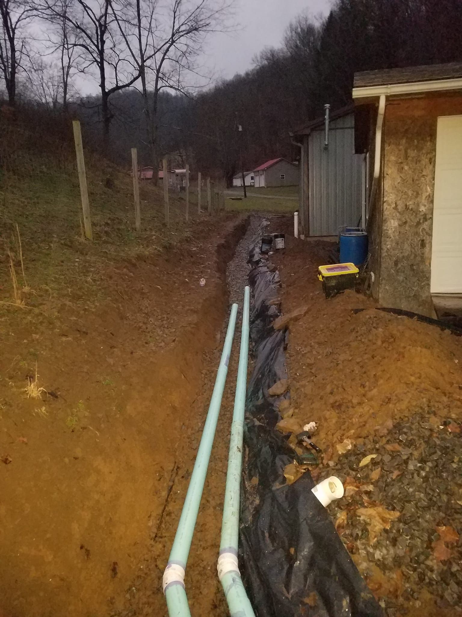 Pipes laid in a trench next to a building and fence, with a hill and cloudy sky in the background.