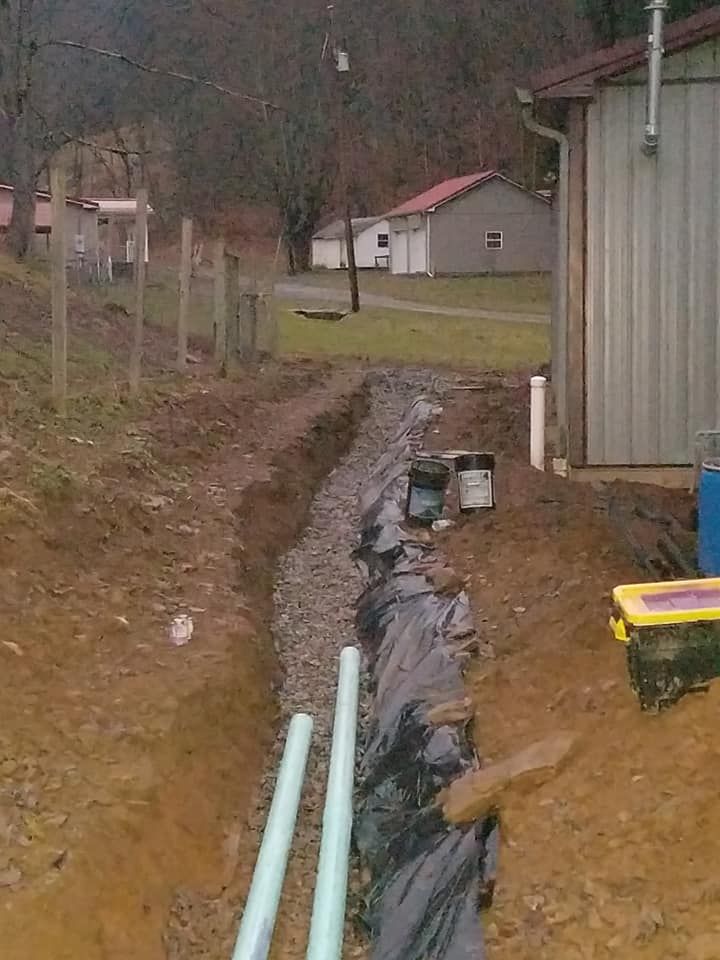 A drainage ditch with gravel, black fabric, and exposed pipes. Buildings and a fence are in the background.