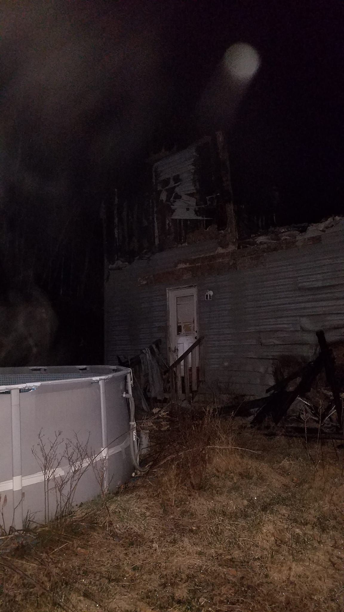 Night scene of a structure, possibly a building, damaged by fire. An above ground pool sits in the yard.